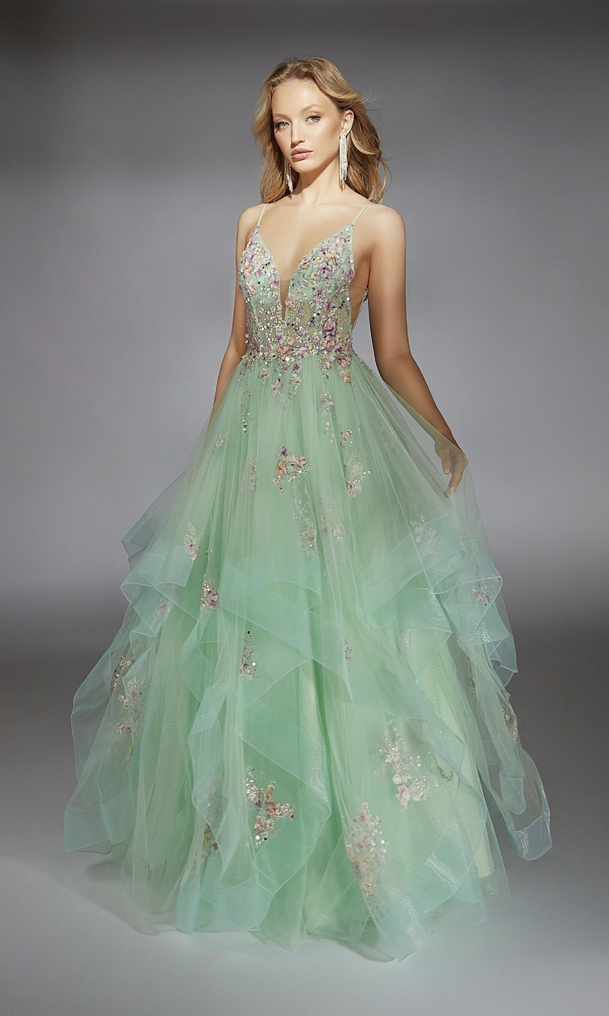A woman poses against a gray background in the Alyce 61735 long prom dress, featuring light green layered tulle, floral embellishments, a deep V-neckline, and thin straps.